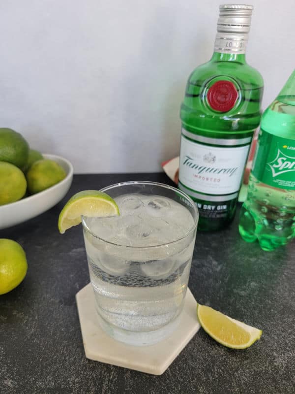 Gin and Sprite in a cocktail glass on a coaster next to a bowl of limes, bottle of gin, and bottle of Sprite