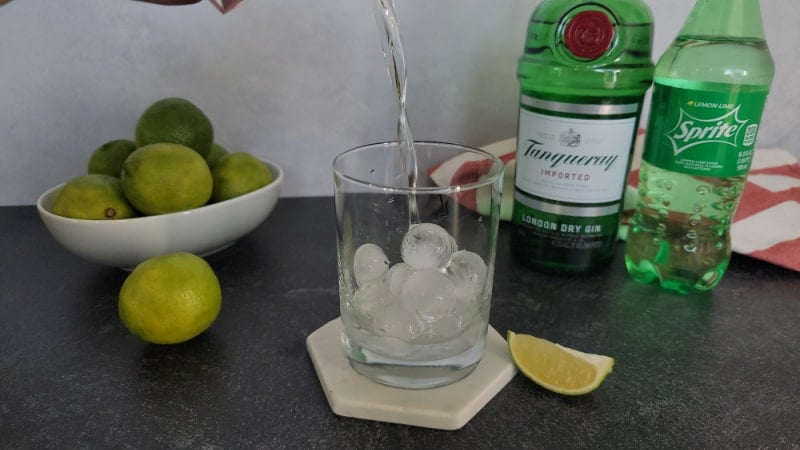 Clear liquid pouring into cocktail glass next to a bowl of limes, bottle of gin, and Sprite