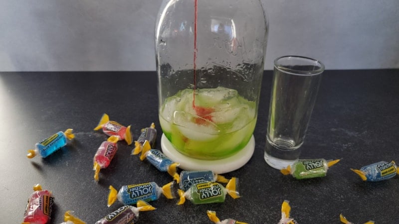 grenadine pouring into jolly rancher shot in cocktail shaker surrounded by jolly rancher candies