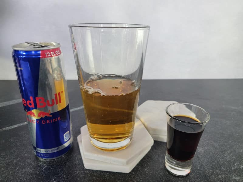 Red Bull can next to a tall glass half full of Red Bull and a shot of Jager for a Jägermeister Bomb Cocktail