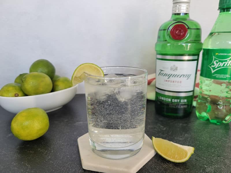 Gin and Sprite garnished with a lime next to a bottle of gin, sprite, and a bowl of limes