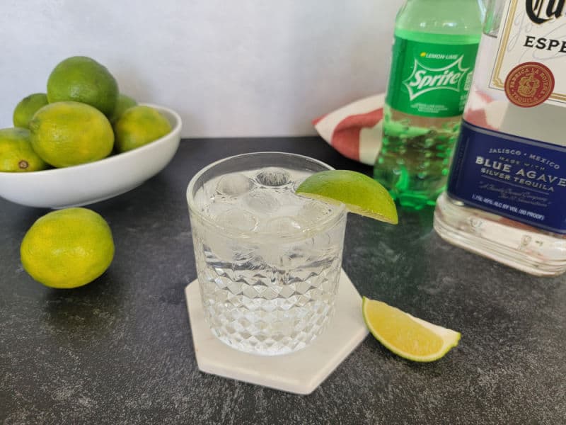 Tequila and Sprite cocktail in a rocks glass next to a bowl of limes, bottle of Sprite, and Bottle of tequila