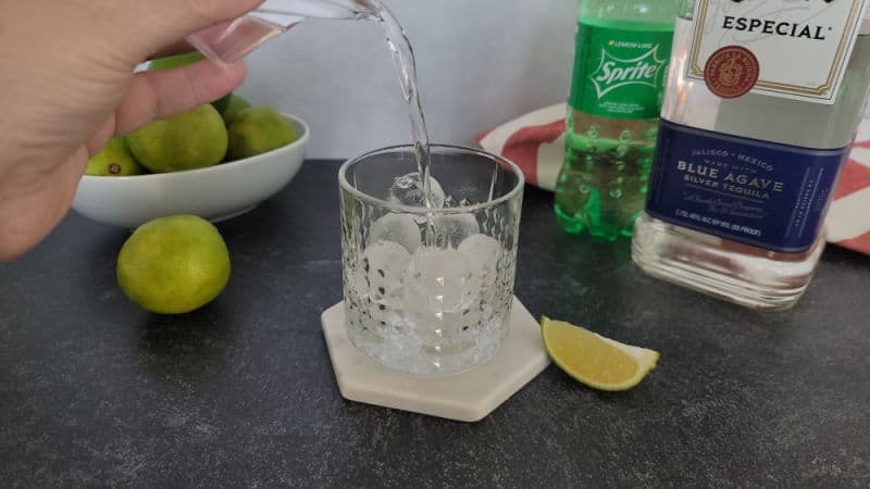 Tequila pouring into a rocks glass next to a bowl of limes, bottle of sprite, and bottle of blue agave tequila