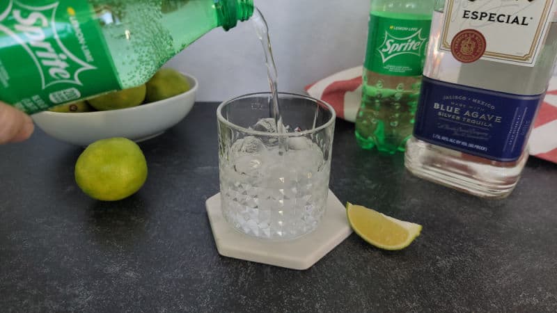 Sprite pouring into a rocks glass next to a bowl of limes, a bottle of Sprite, and a bottle of tequila