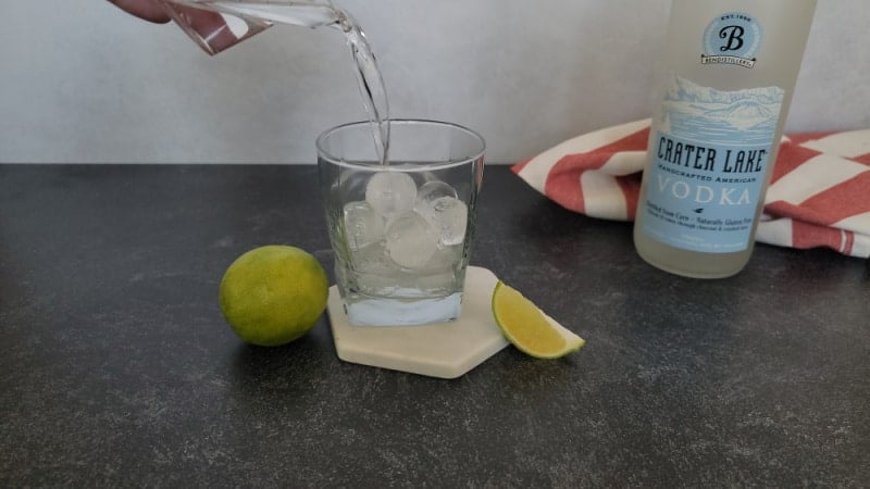 Vodka pouring into a cocktail glass with ice cubes next to a bottle of Crater Lake Vodka