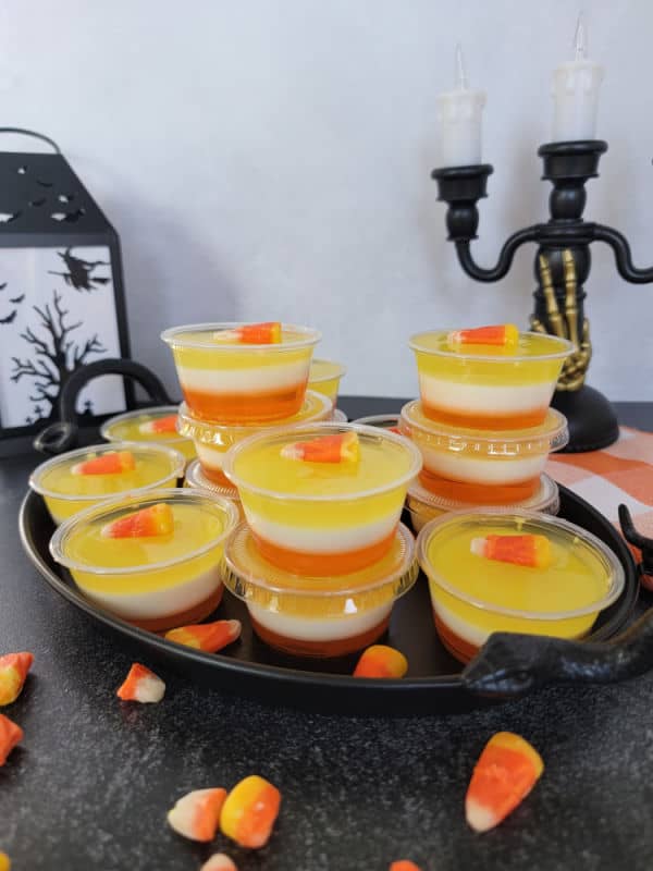 Layered Candy Corn Jello Shots in plastic cups on a black tray next to a lantern and candy corn candies