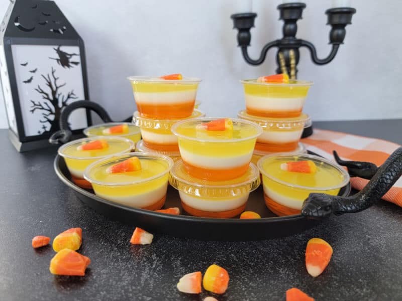 Layered Candy Corn Jello Shots in plastic cups on a black tray next to a lantern and candy corn candies