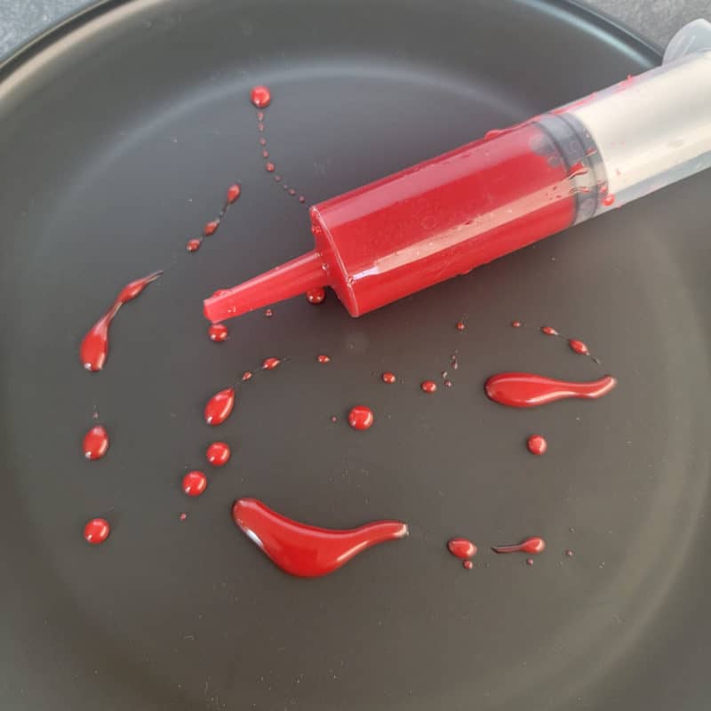 Edible Blood in a plastic syringe on a black plate with edible blood splatters