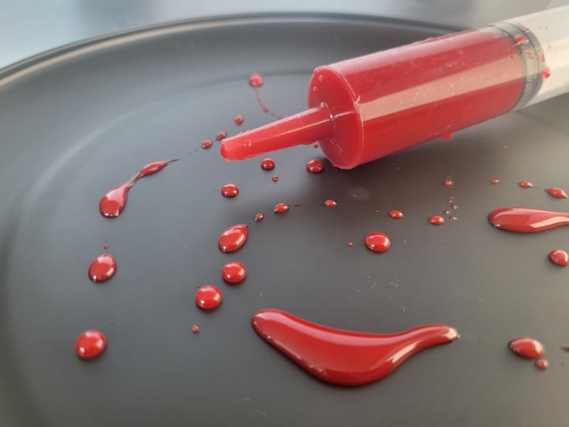 Edible Blood in a plastic syringe on a black plate with edible blood splatters
