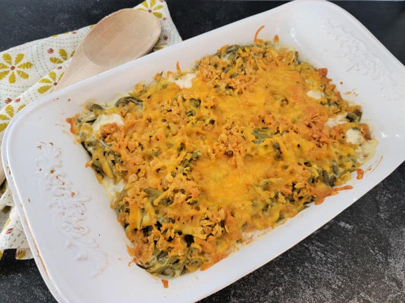 Cheesy green bean casserole in a white casserole dish with a wooden spoon and cloth napkin
