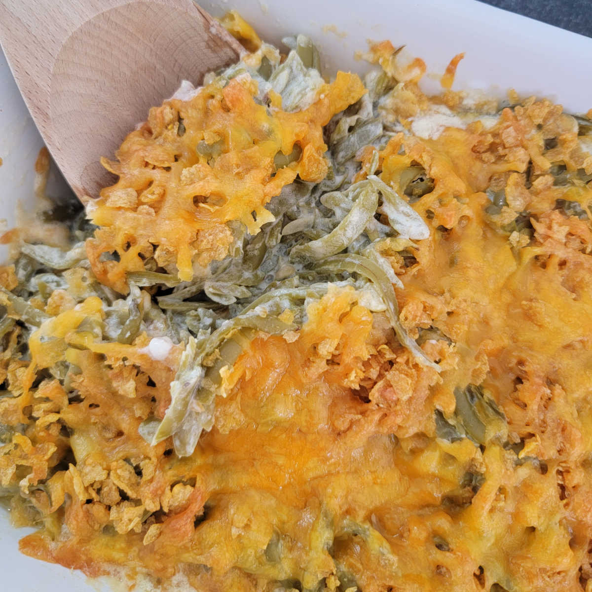 wooden spoon scooping cheesy green beans from a white casserole dish