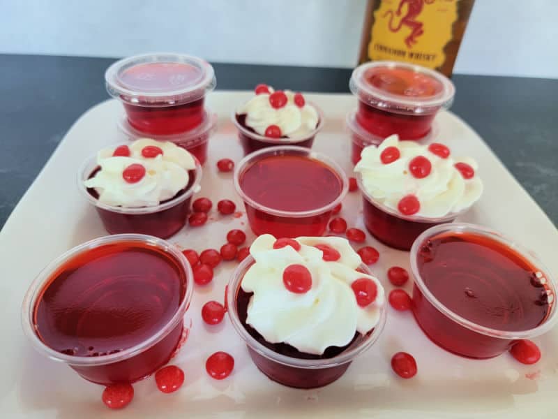 Fireball Jello Shots in plastic cups some garnished with whipped cream and red hots with a bottle of Fireball Cinnamon Whiskey