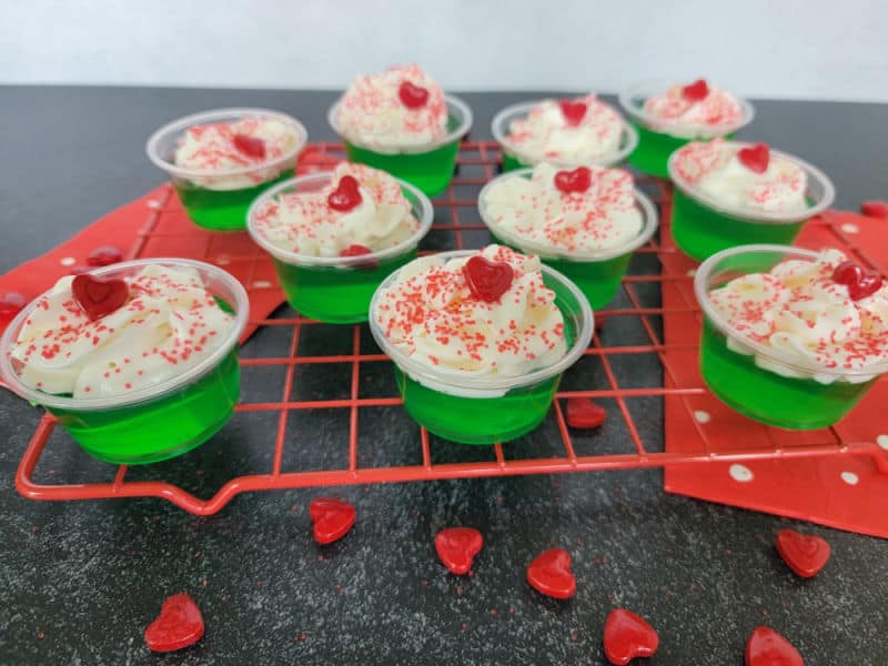 Grinch Jello Shots garnished with whipped cream, red sprinkles, and a red candy heart sitting on a red rack