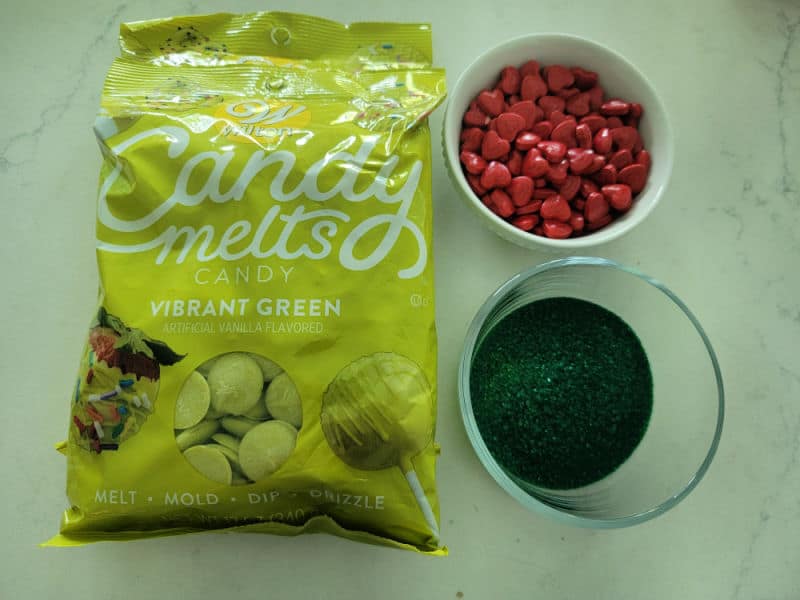 Wilton Green Candy Melts in the bag next to a bowl of red candy hearts and a bowl of green sprinkles 