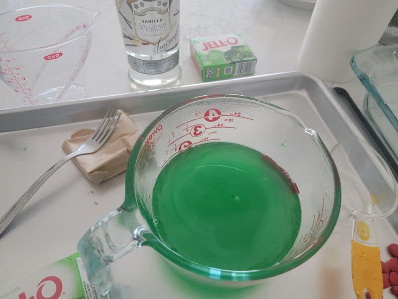 green jello in a large measuring bowl on a baking sheet with vodka, and a fork for Grinch Jello Shots