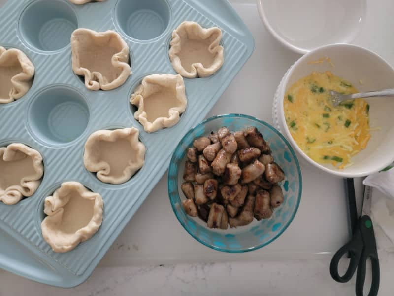 biscuit cups in a blue muffin tin next to a bowl of sausage and a bowl of eggs, cheese, and green onions on a white counter