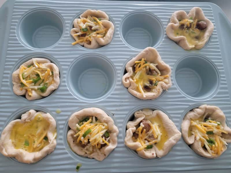 Sausage Breakfast Biscuit Cups in a blue muffin tin before baking. Eggs, cheese, green onion, and sausage filled in the biscuit cups