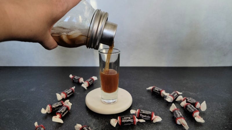 Pouring a tootsie roll shot from a cocktail shaker into a shot glass surrounded by tootsie rolls