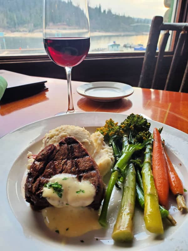 steak and vegetables on a white plate with a glass of wine and view of Bass Lake at Duceys on the Lake