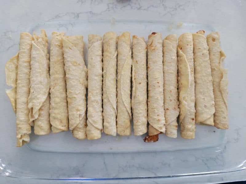 Frozen taquitos spread in a glass baking dish for Taquito enchiladas