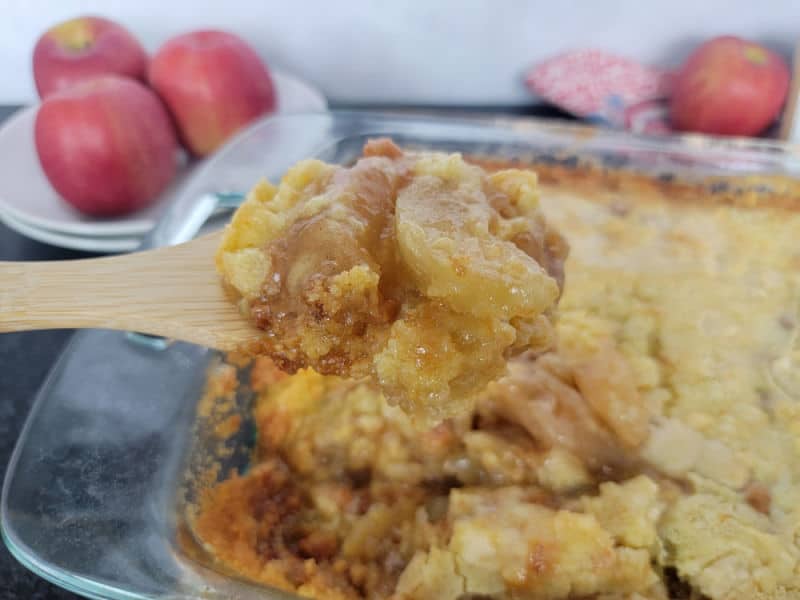 Wooden spoon scooping caramel apple dump cake from a glass casserole dish with apples in the background