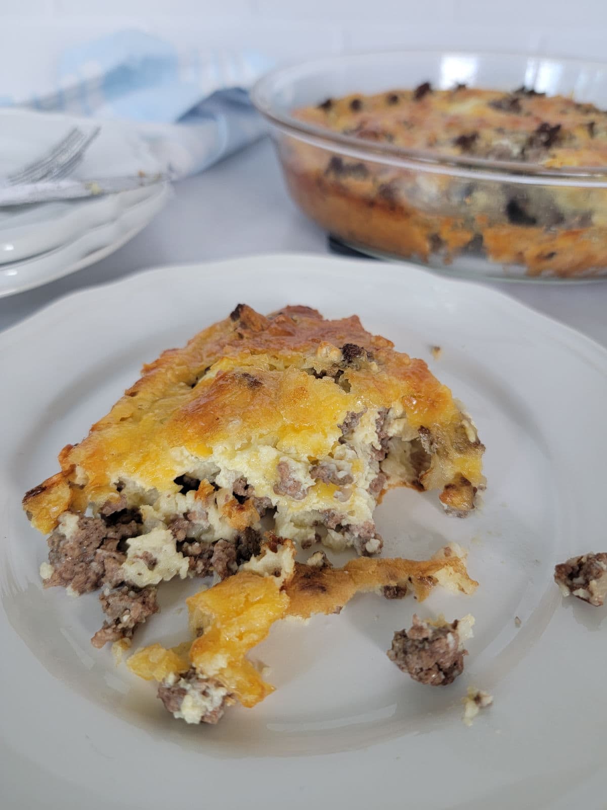 Slice of impossible pie on a white plate with the baking dish behind it and a pile of plates