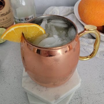 Orange Moscow Mule in a copper mule mug next to a bowl of oranges and ginger beer and vodka