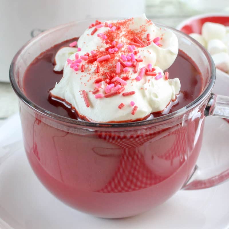 Red Velvet Hot Chocolate in a glass mug topped with whipped cream and pink sprinkles