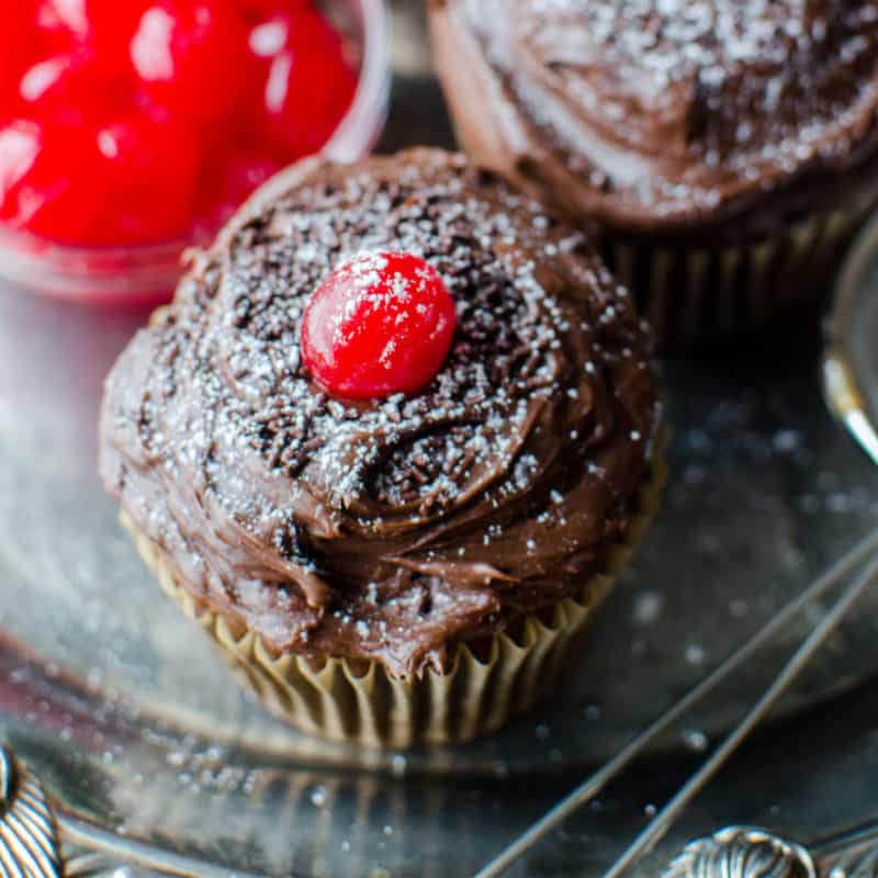 Black Forest Cupcake garnished with a maraschino cherry on a silver platter by a bowl of cherries