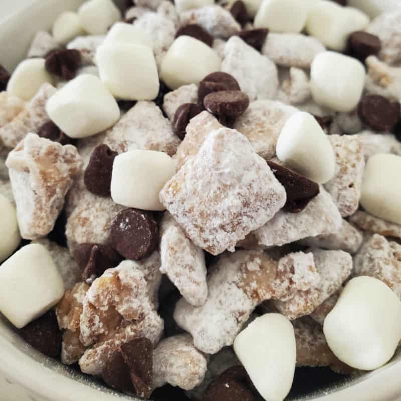 Hot chocolate muddy buddies piled in a white bowl with mini marshmallows and chocolate chips