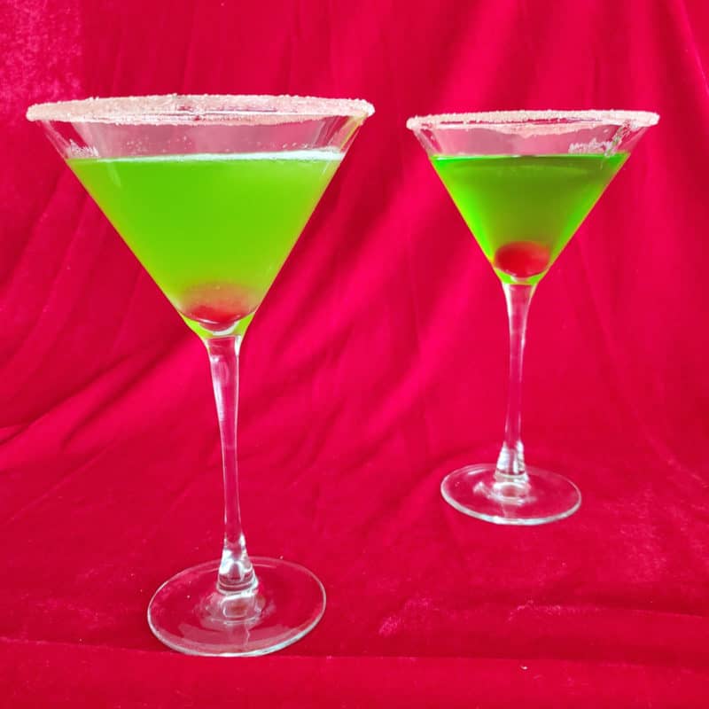 Two Bright green cocktails with cherries in them in martini glasses with a sugared rim.