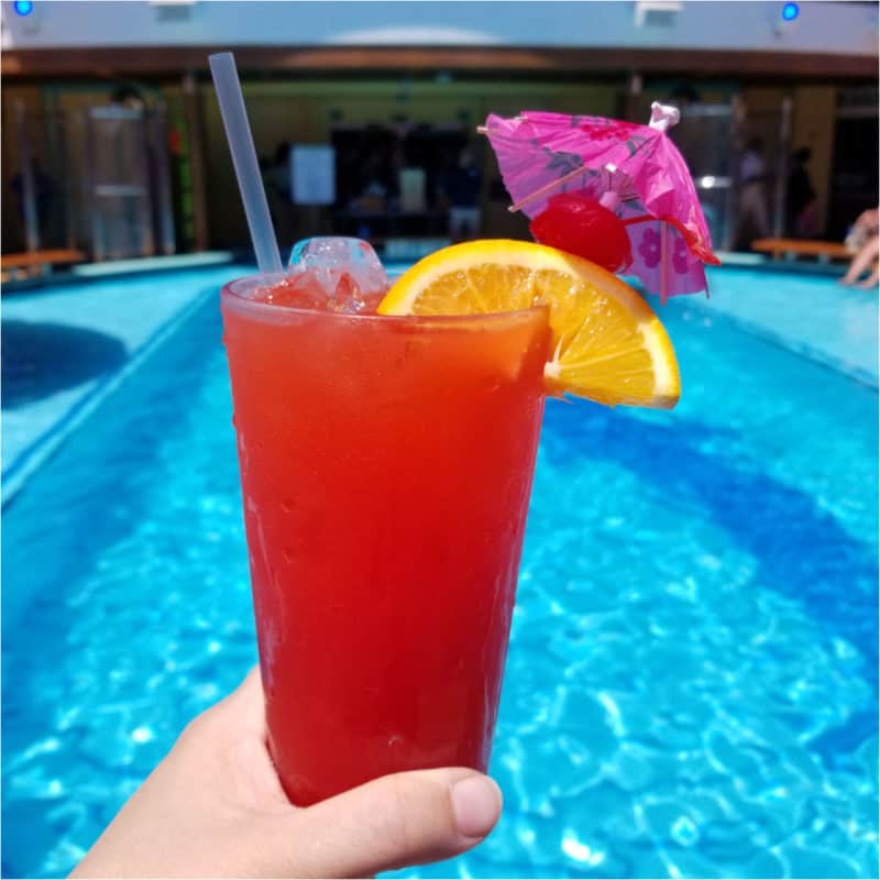hand holding a red cocktail with orange wheel and pink umbrella by the pool