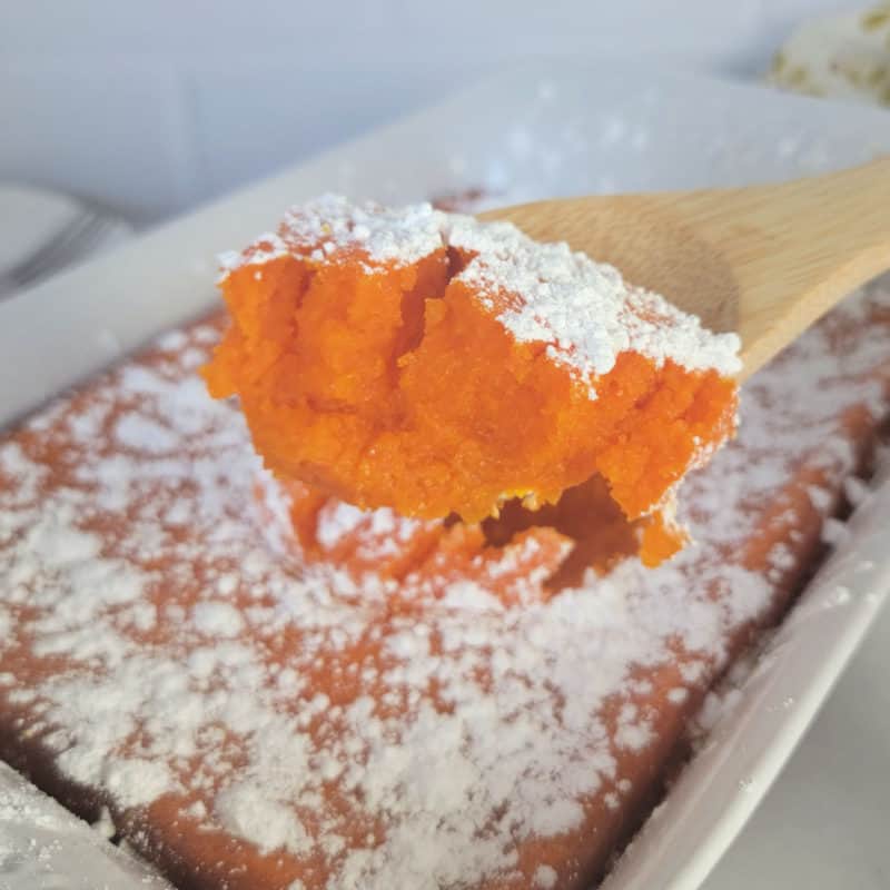 carrot souffle with powdered sugar garnish on a wooden spoon above a white casserole dish