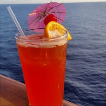 red cocktail with an orange wheel, cherry and pink umbrella with the ocean behind it