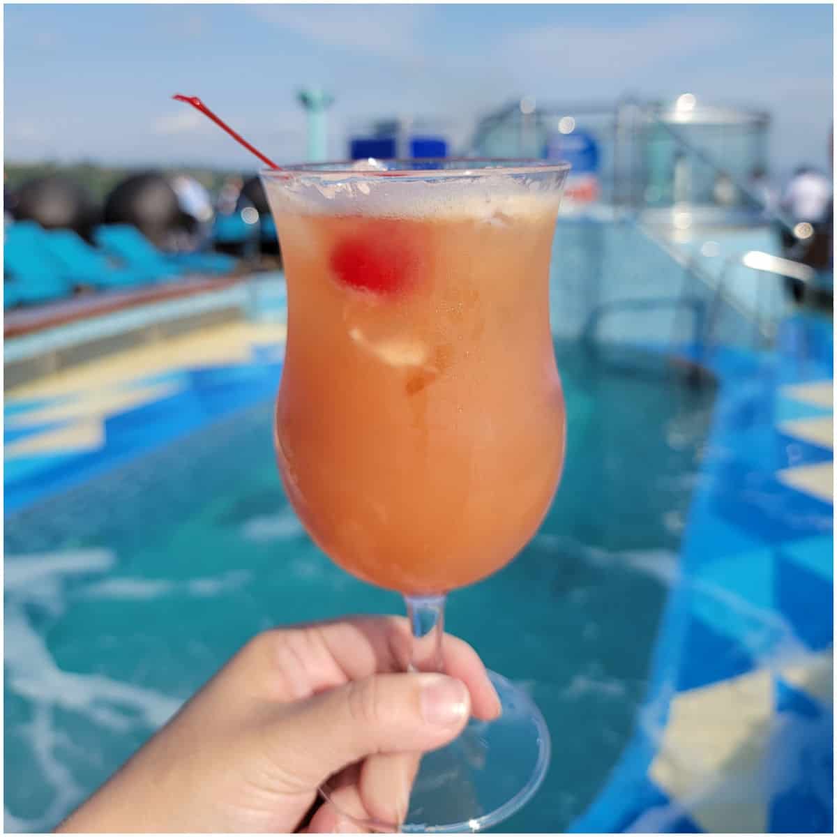 pink orange cocktail in a hurricane glass with a cherry in front of a pool