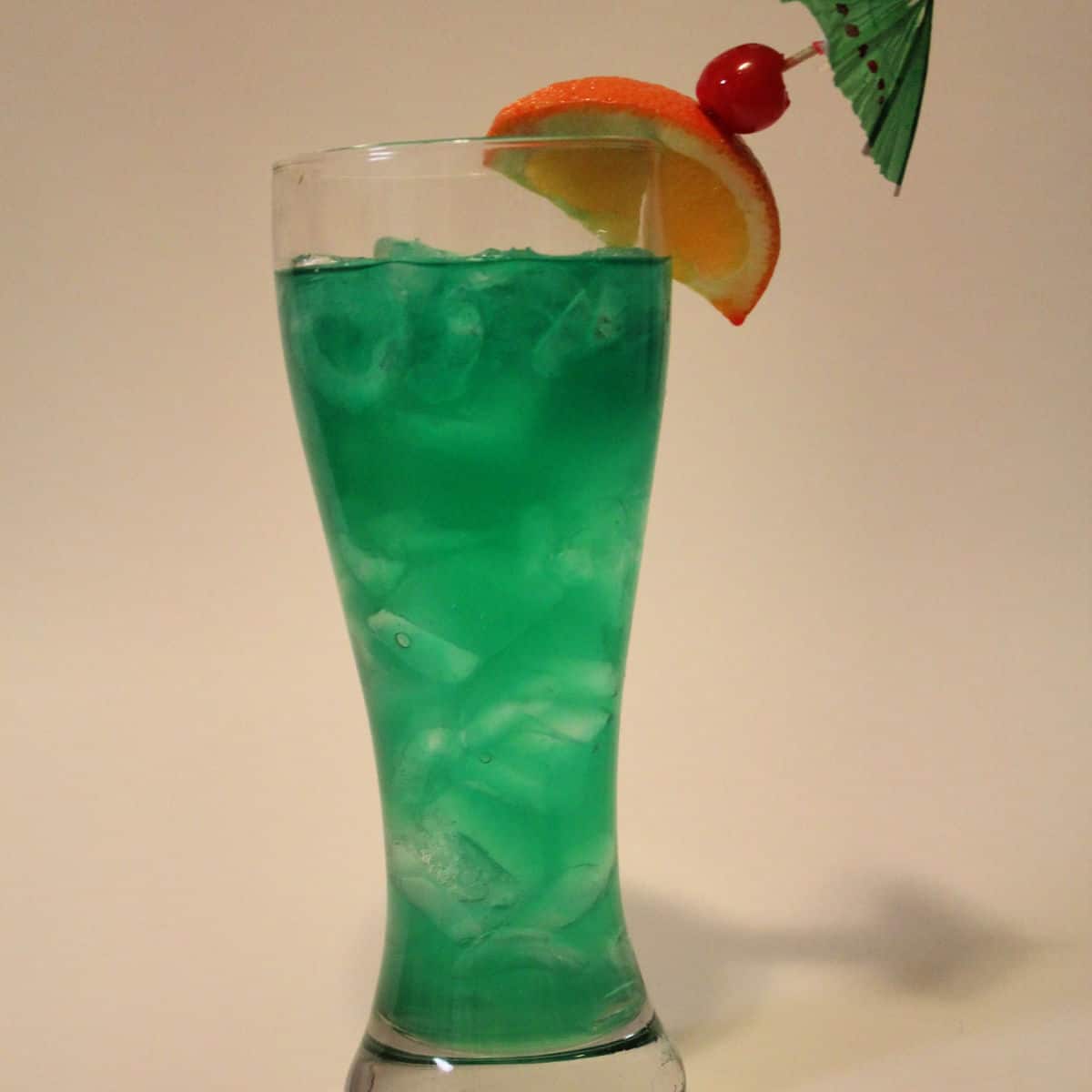 Bright green cocktail in a tall glass with orange slice and cherry garnish