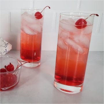 Two red cocktails in tall glasses garnished with maraschino cherries next to a bowl of cherries
