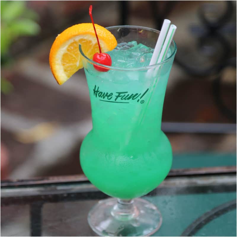 green cocktail in a hurricane glass with orange wedge and maraschino cherry garnish