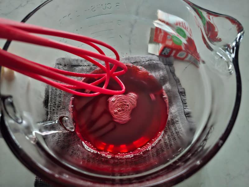 strawberry jello mix in a glass bowl with a red whisk