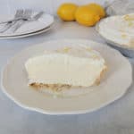 Slice of lemon ice box pie on a white plate with lemons and the full pie behind it