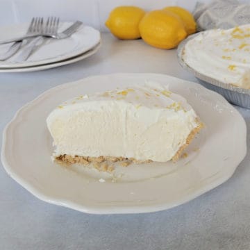 Slice of lemon ice box pie on a white plate with lemons and the full pie behind it