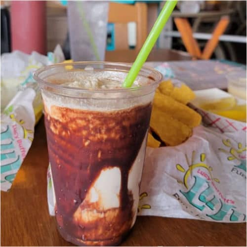 Chocolate bushwacker in a plastic cup with a green straw, lulu's chips and salsa next to the glass