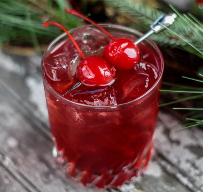 red cherry cocktail with two maraschino cherries for garnish next to greenery
