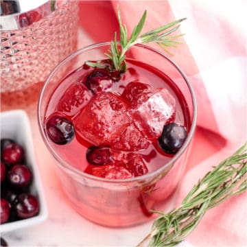 red cocktail with cranberries and rosemary next to a bowl of cranberries and a glass cocktail shaker