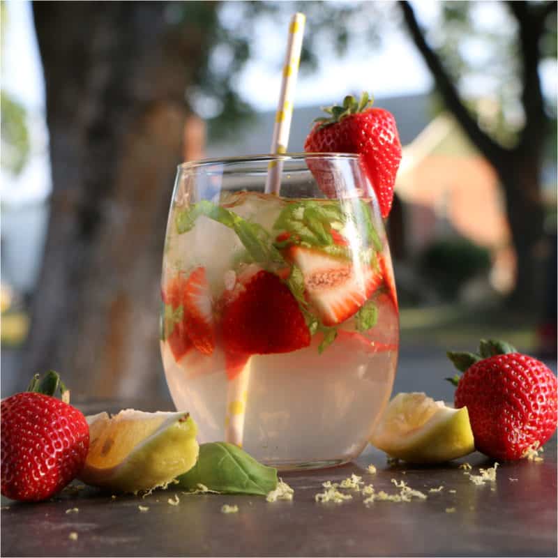 strawberries and basil in a lemonade cocktail with paper straw next to more strawberries and a lemon wedge