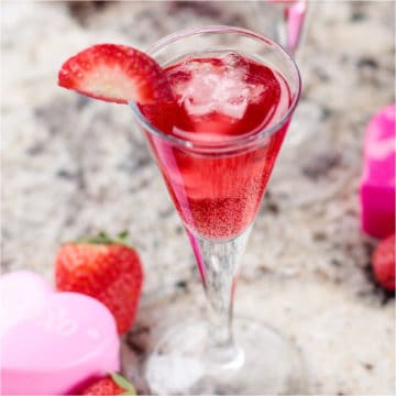 red champagne cocktail garnished with a strawberry next to pink hearts and strawberries