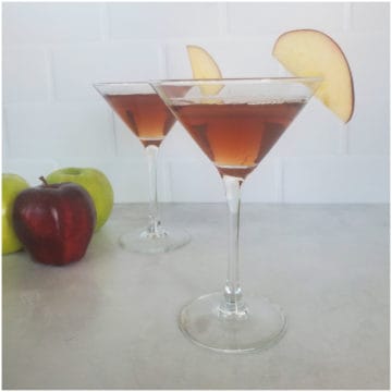 Two red brown martinis with apple slices in a martini glass next to green and red apples