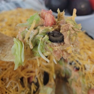 Tortilla holding 7 layer taco dip above a full casserole dish