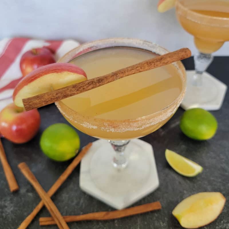 Apple Cider Margarita with a cinnamon stick surrounded by apples and lime wedges