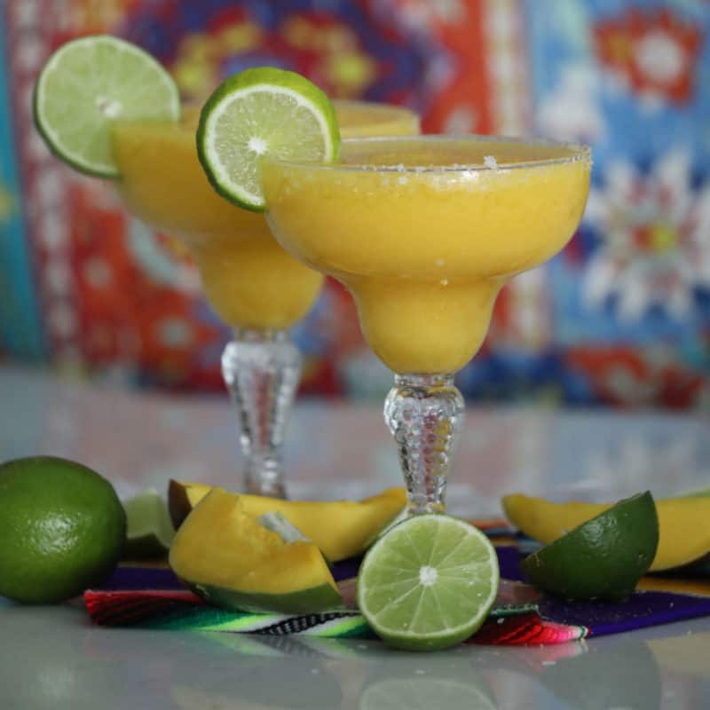 Two mango margaritas in margarita glasses with mangos and limes 
